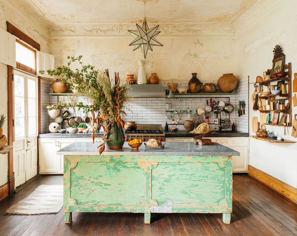 keukengedeelte met groot rustiek eiland in het midden met groen afbladderend hout en open planken aan de achterste muur met enkele grote houten schalen en kannen en aan de rechterkant planken met boeken en aan de tegenoverliggende muur een kleine dubbele deur die naar buiten leidt